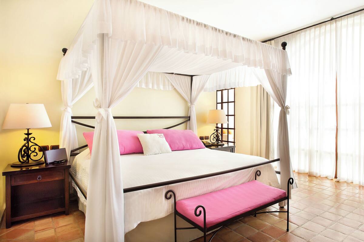 A four-poster canopy bed with a bench, bedside tables with lamps, and a pink armchair.