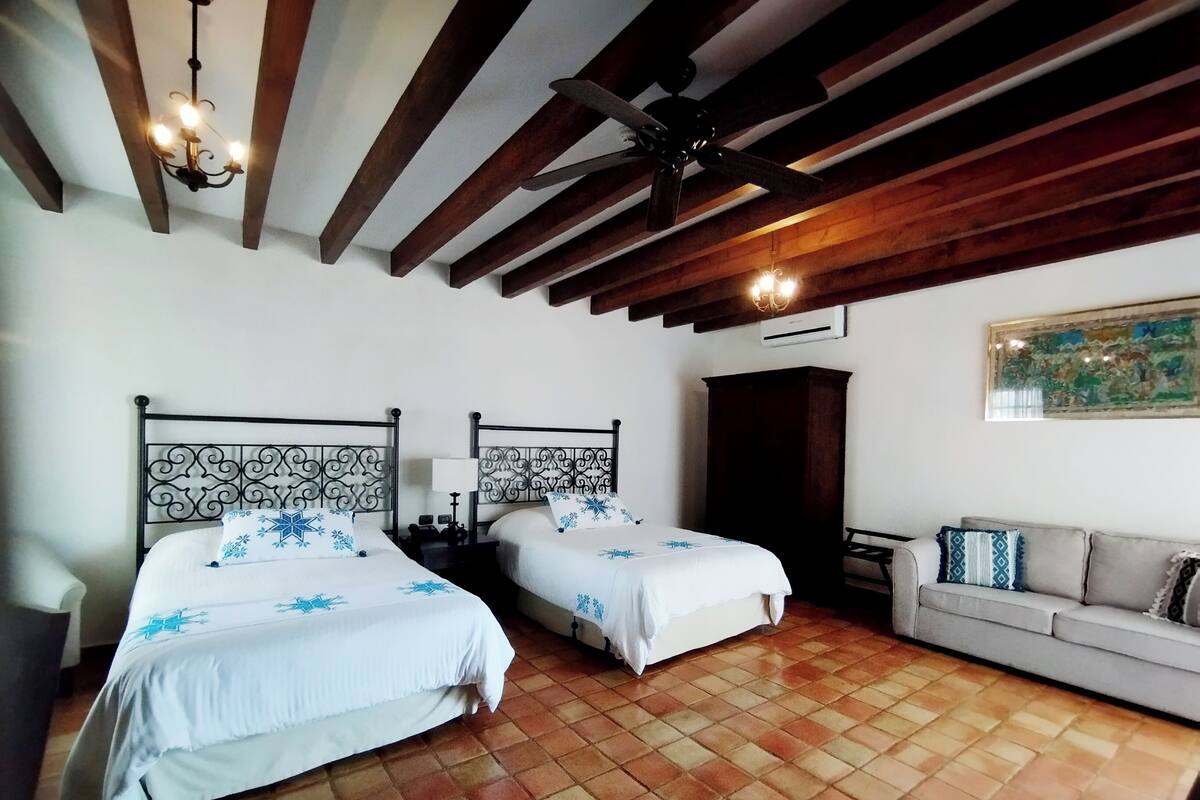 A hotel room with two beds, a sofa, a ceiling fan, and a painting on the wall.