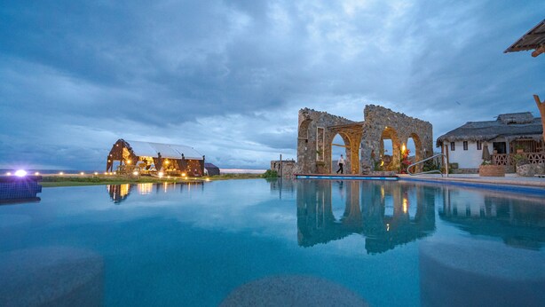 A tranquil infinity pool reflecting a historic stone structure and a lit building at dusk.