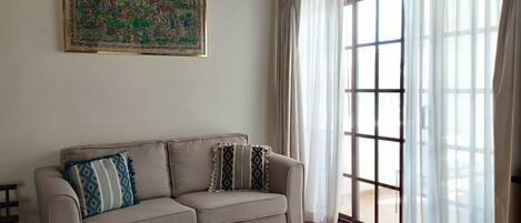 A living room with a beige sofa, a colorful tapestry, and a large window with sheer curtains.