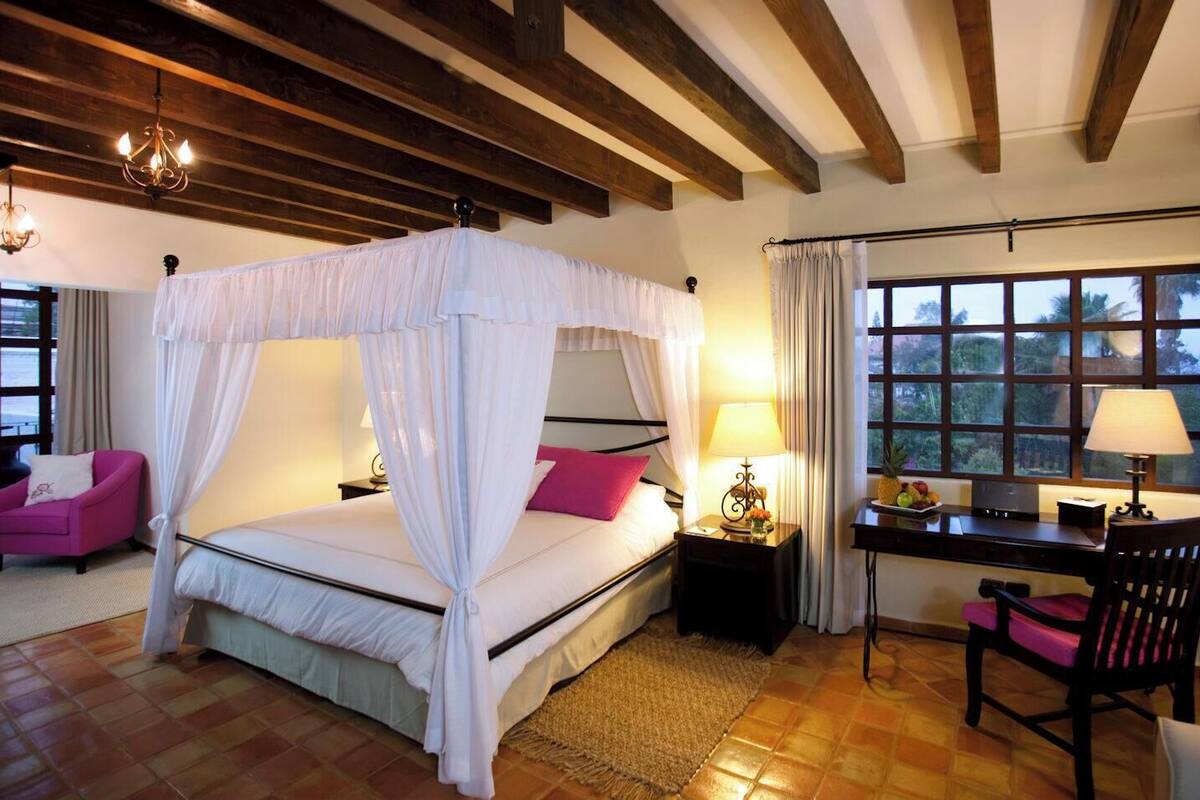 A bedroom with a four-poster bed, a desk with a chair, and a view of the outdoors through large windows.