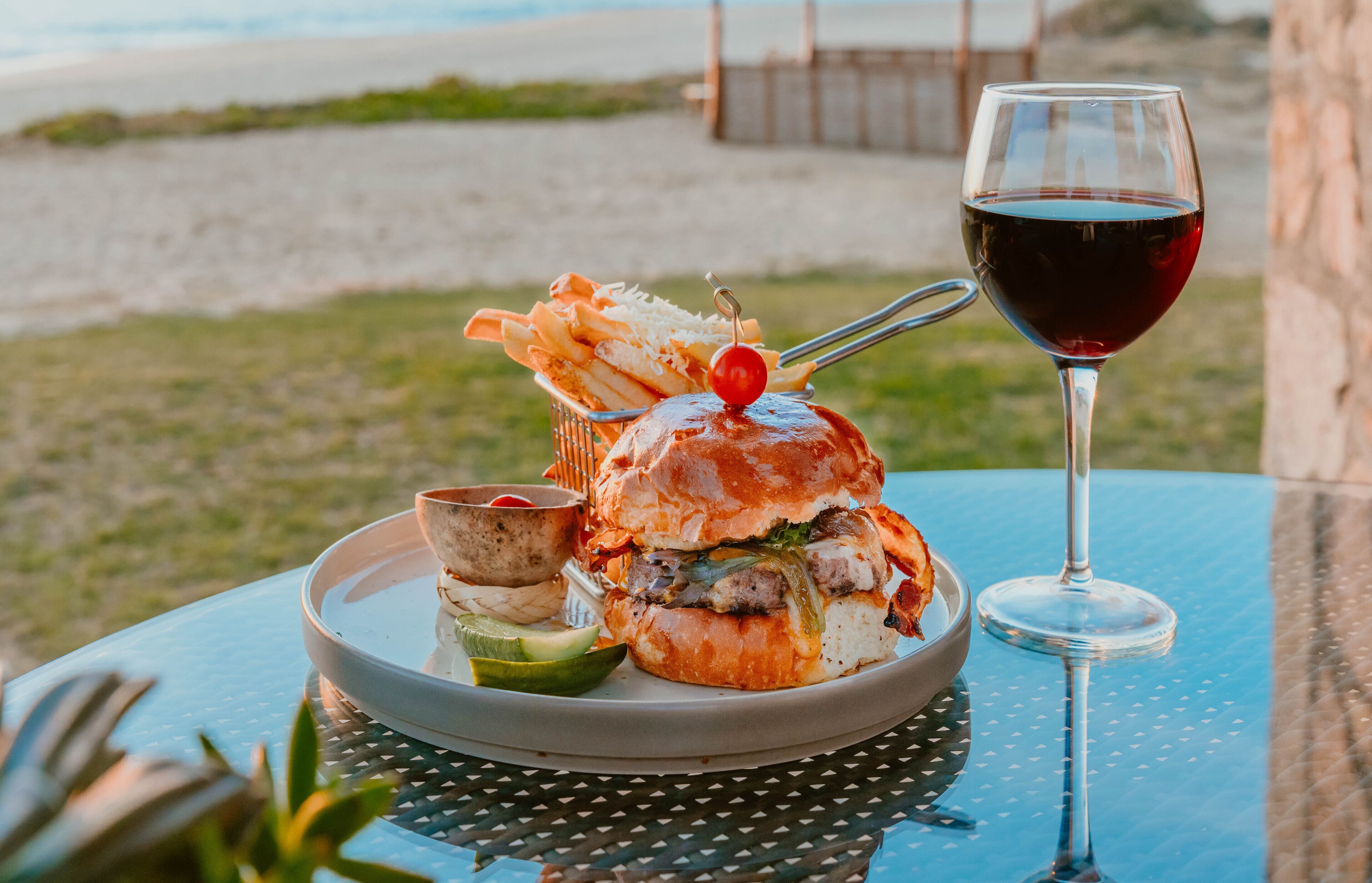 A meal consisting of a burger, fries, and a glass of red wine on an outdoor table with a view of the beach.