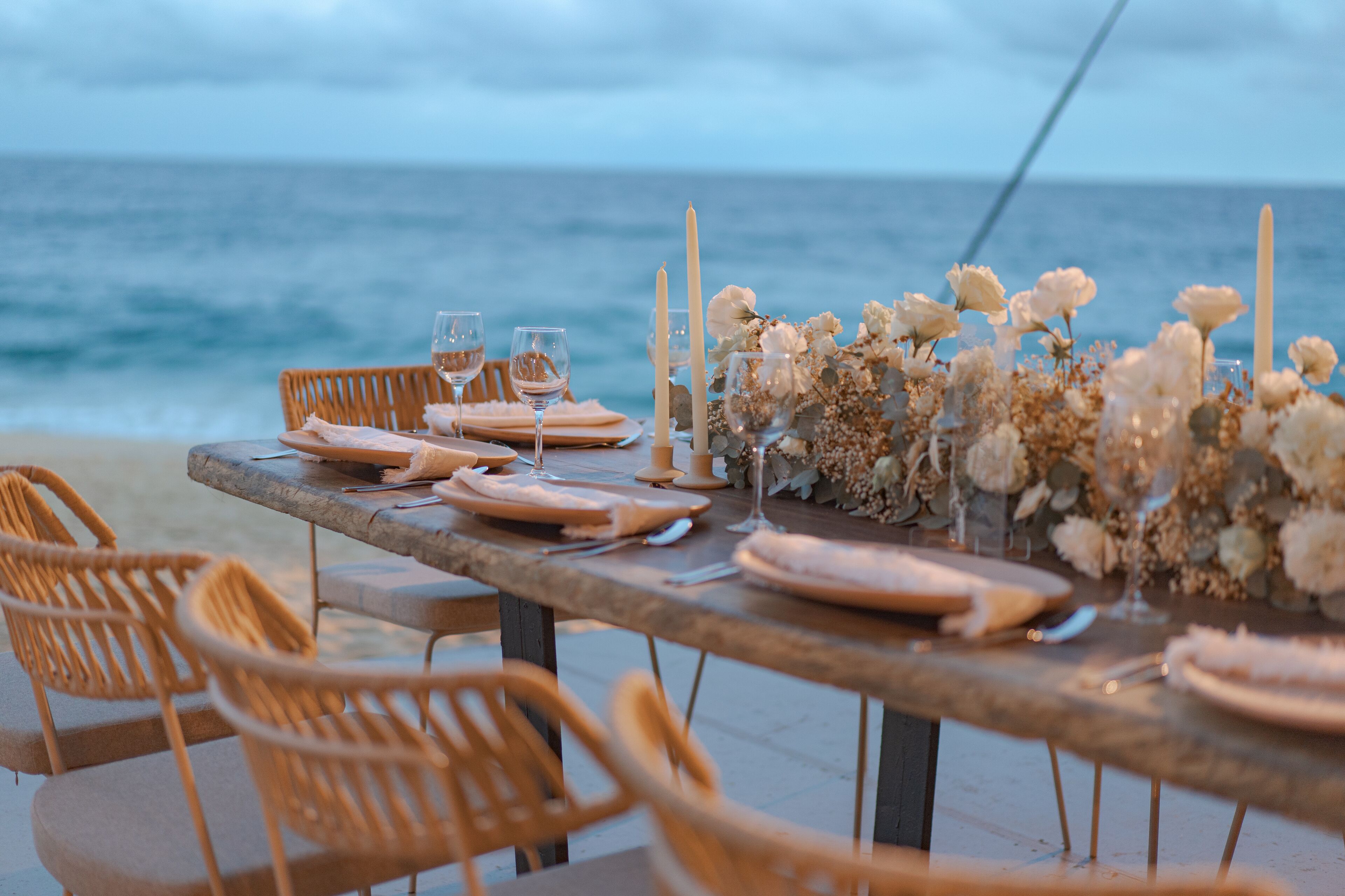 A table set for a meal with chairs, candles, and flowers by the ocean.