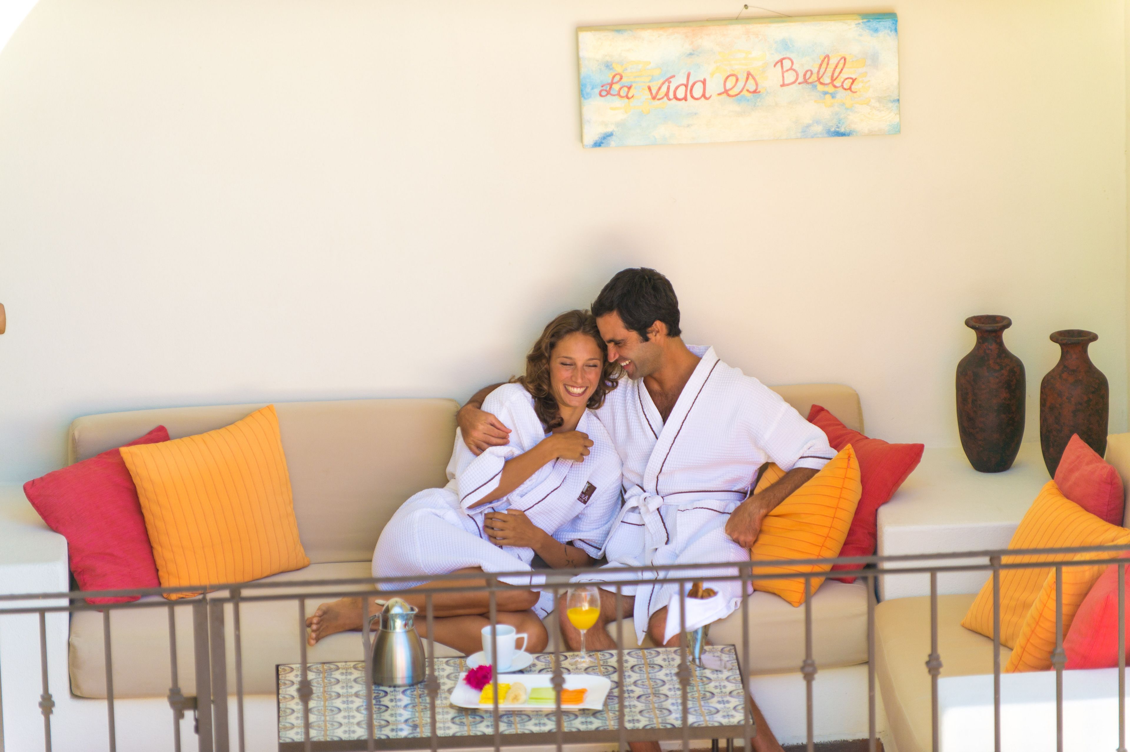 A couple in white bathrobes sitting on a beige sofa with orange pillows, a table with a teapot, cups, and a fruit plate in front of them.