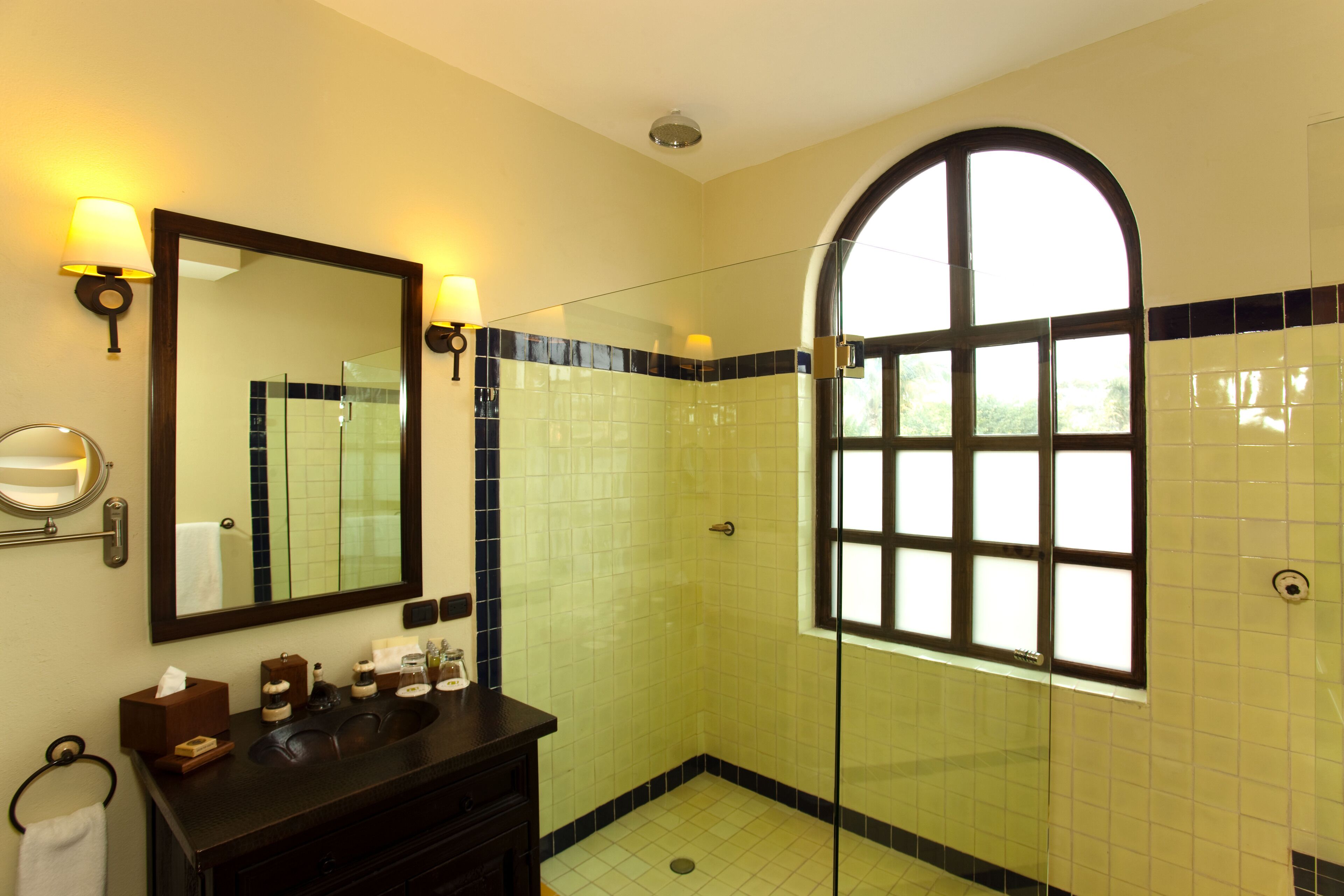 A bathroom with a large mirror, two wall-mounted lamps, a dark wooden vanity, a glass-enclosed shower, and a window with a view of greenery.