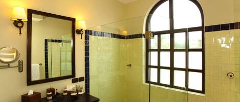 A bathroom with a large mirror, two wall-mounted lamps, a dark wooden vanity, a glass-enclosed shower, and a window with a view of greenery.