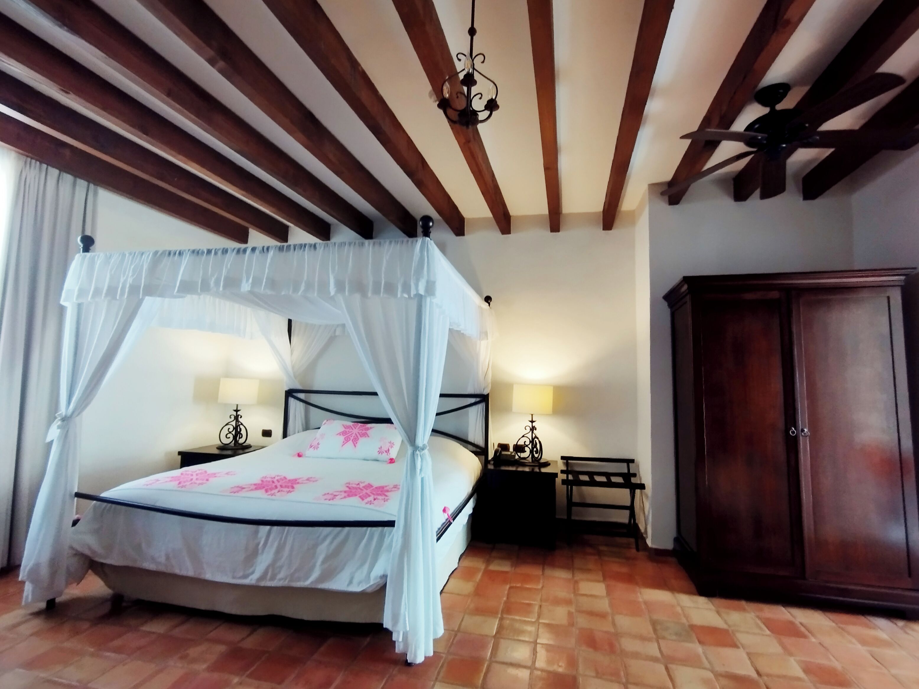 Une chambre à coucher avec un lit à baldaquin, des poutres en bois au plafond, un ventilateur de plafond, une table de chevet avec une lampe et une armoire en bois.