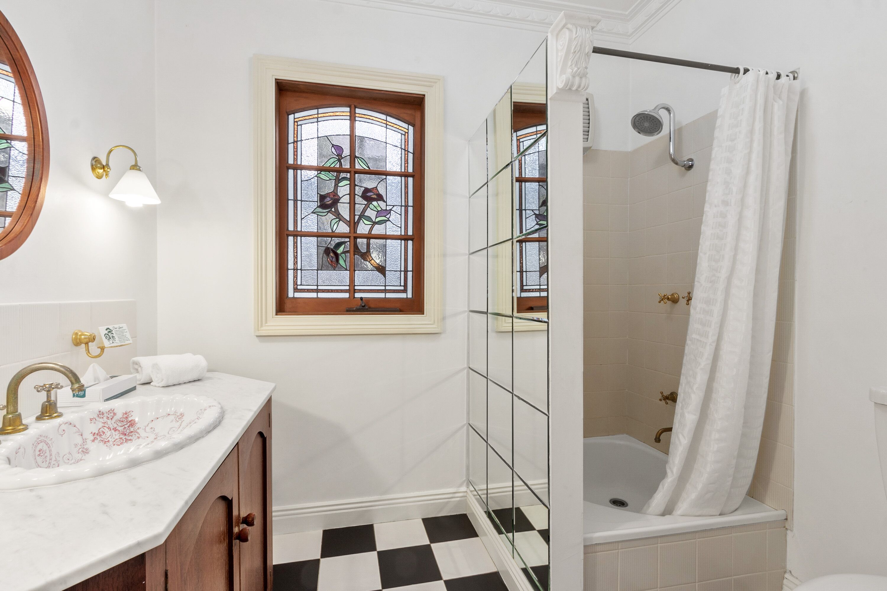 Une salle de bain avec un lavabo blanc, une douche dotée d’une paroi en verre, une fenêtre avec des vitraux et un carrelage à motifs géométriques.