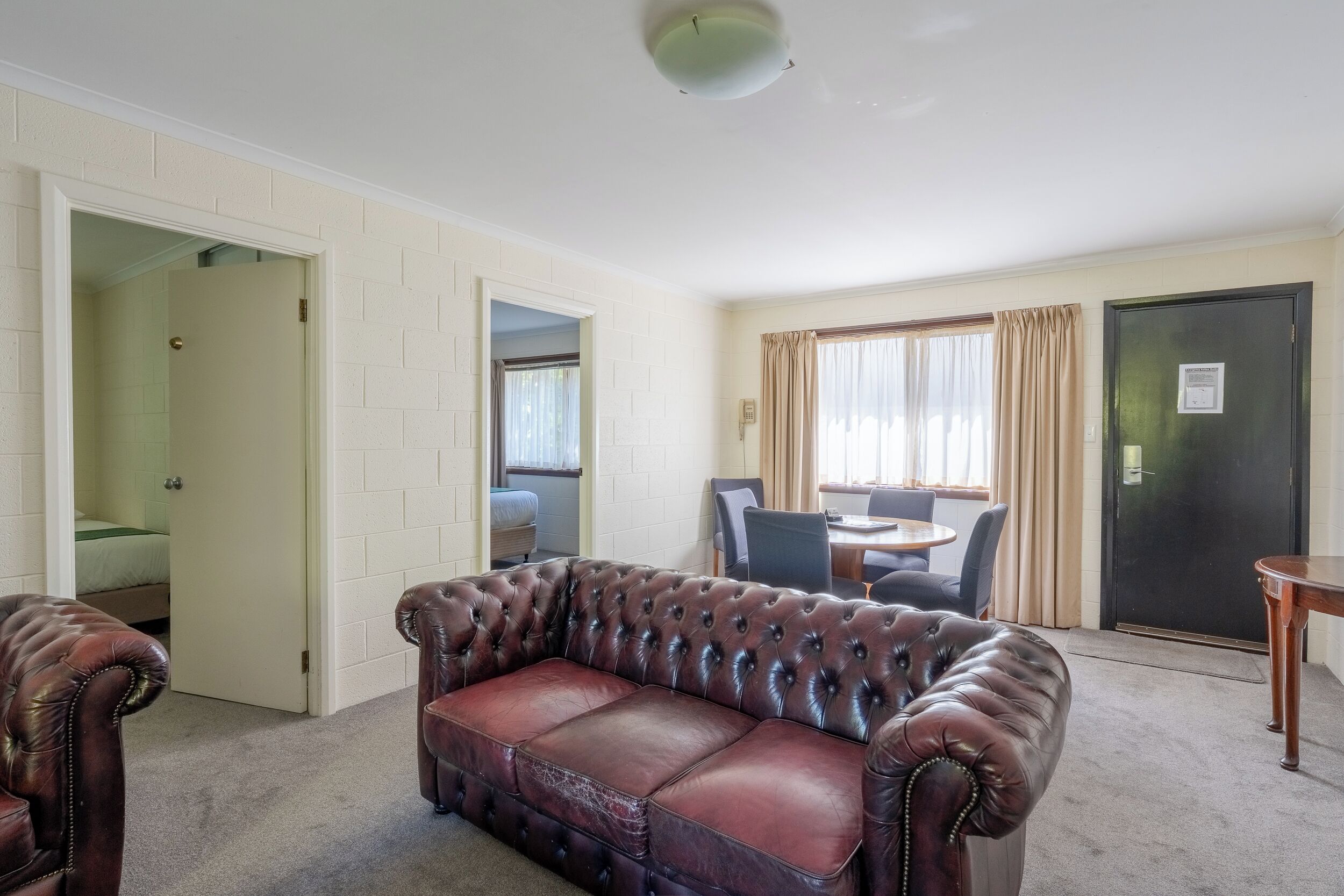 A living room with a leather sofa, a dining table with chairs, and a door leading to a bedroom.