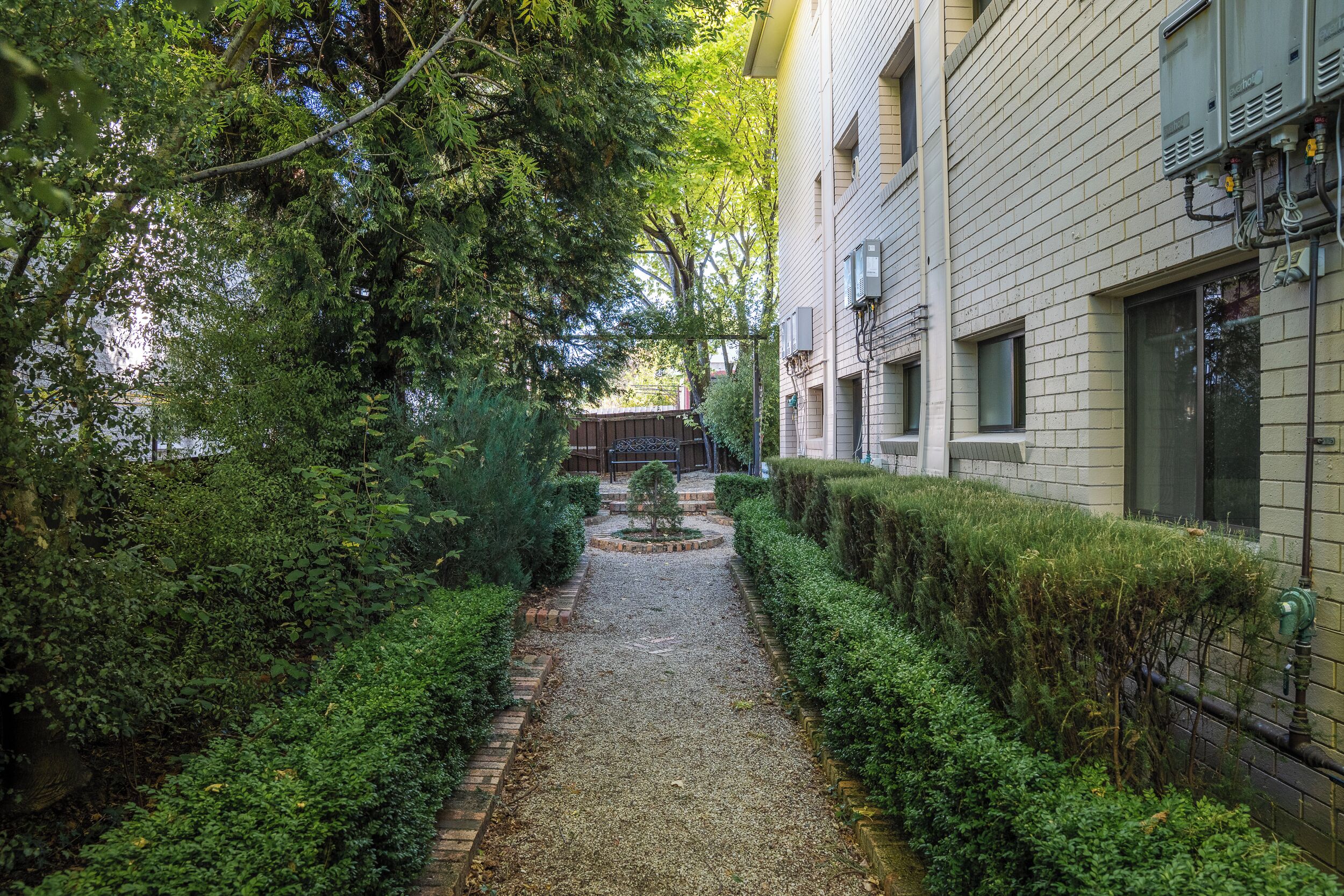 Un sendero estrecho bordeado por setos y árboles cuidadosamente podados, que conduce a un edificio con varias ventanas y cajas eléctricas exteriores.