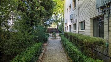Un sendero estrecho bordeado por setos y árboles cuidadosamente podados, que conduce a un edificio con varias ventanas y cajas eléctricas exteriores.