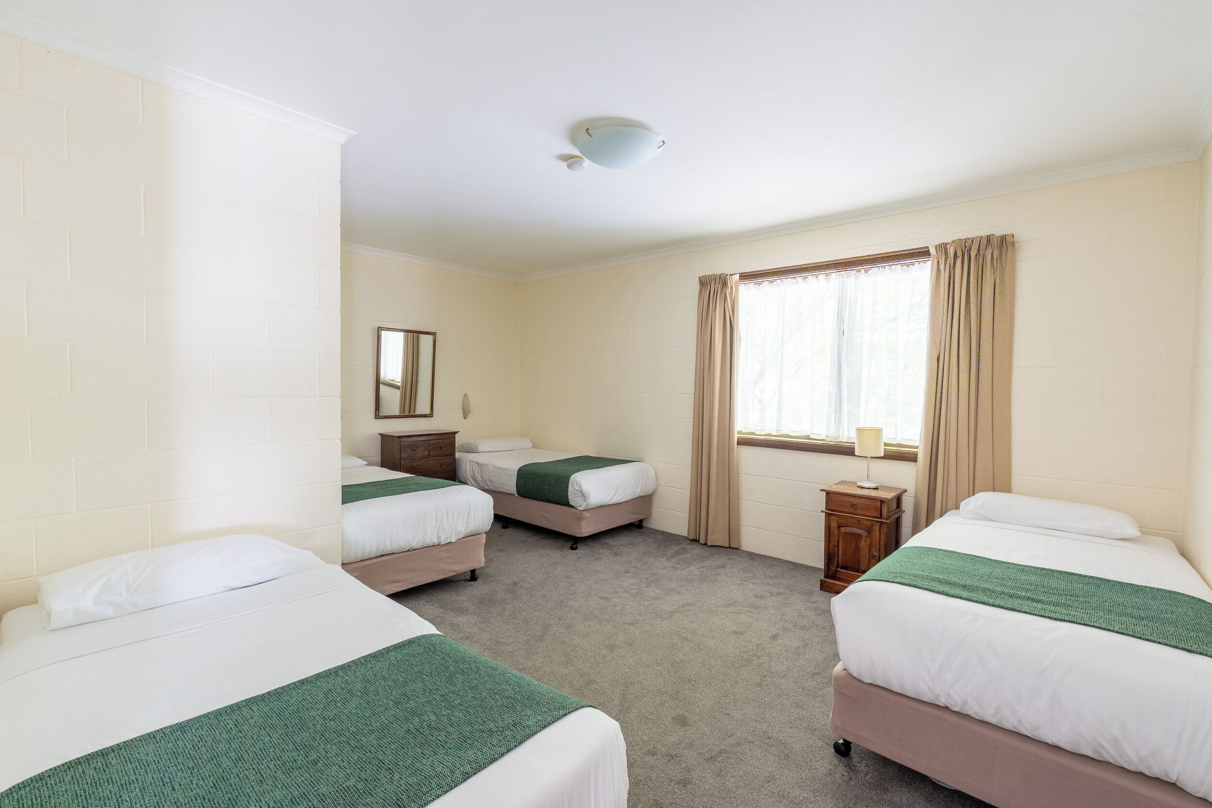 Een hotelkamer met drie eenpersoonsbedden, een klein nachtkastje met een lamp, een raam met gordijnen, en een plafondlamp.