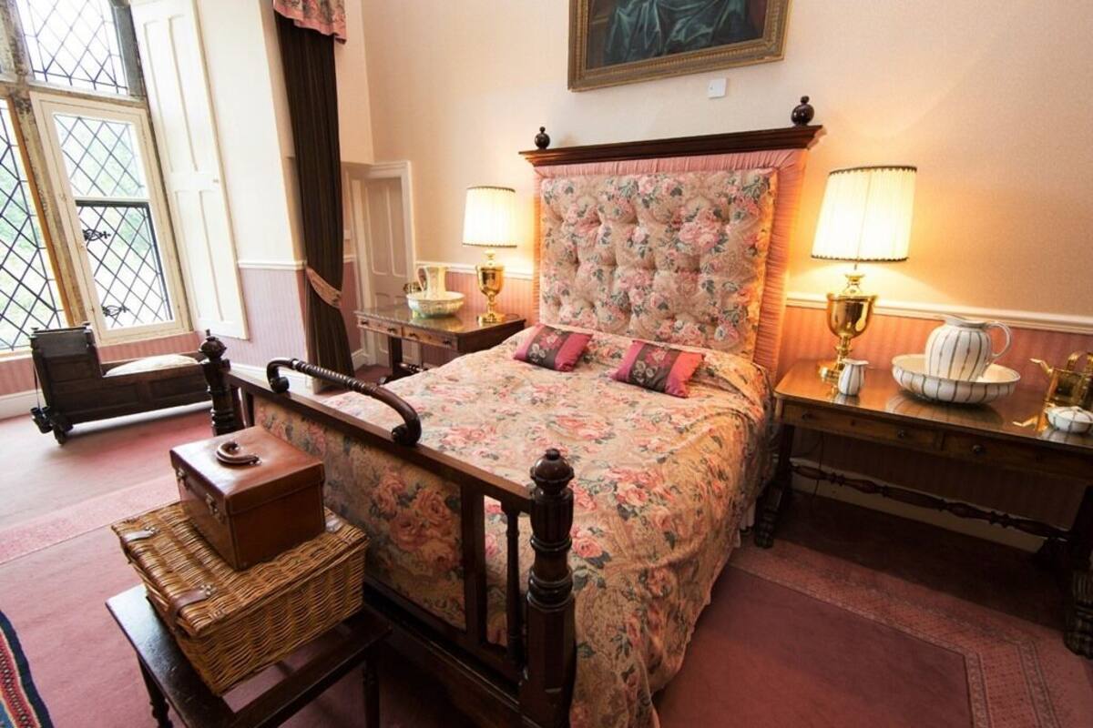 Une chambre avec une couvre-lit à motifs floraux, une tête de lit en bois, deux lampes de chevet, une commode en osier et une vue sur l’extérieur par une fenêtre.