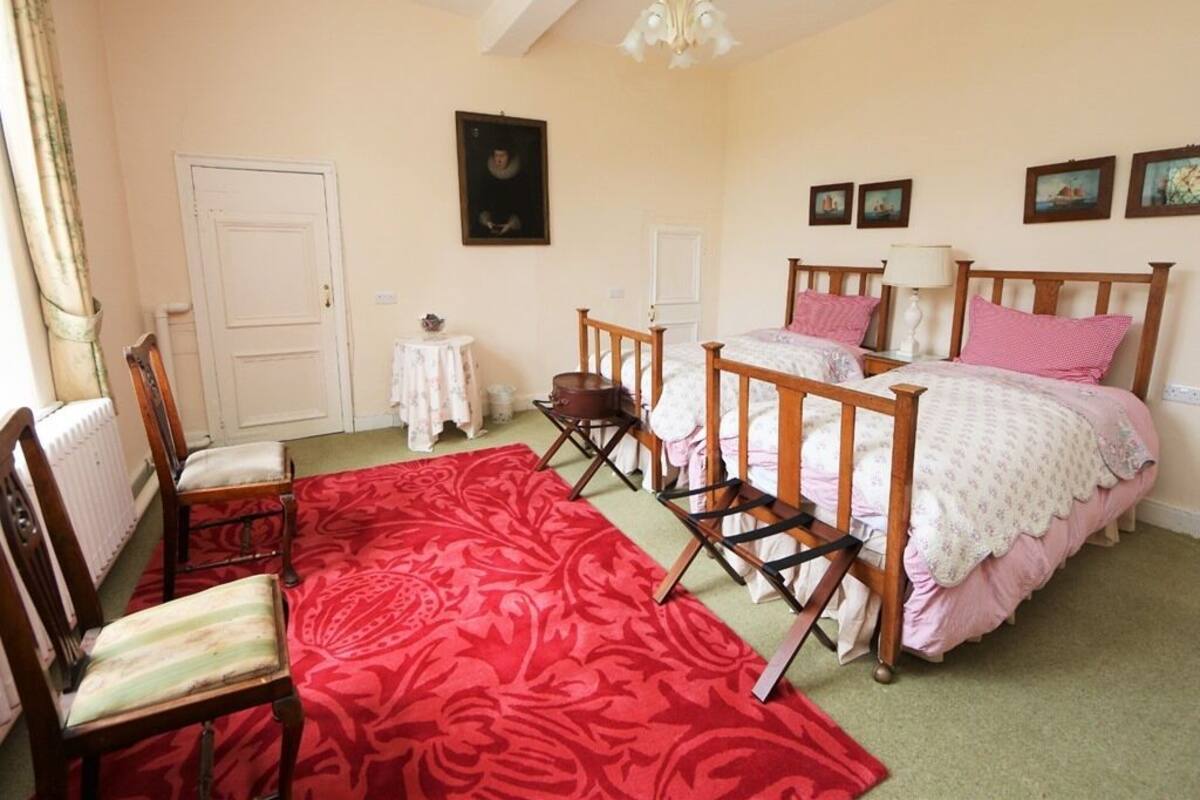 Une chambre avec deux lits, un tapis rouge à motifs, des chaises en bois et un radiateur blanc.