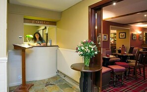 A reception area with a counter, a desk, a vase with flowers, and a dining area with chairs and tables. - The Argyll Hotel (Ullapool)