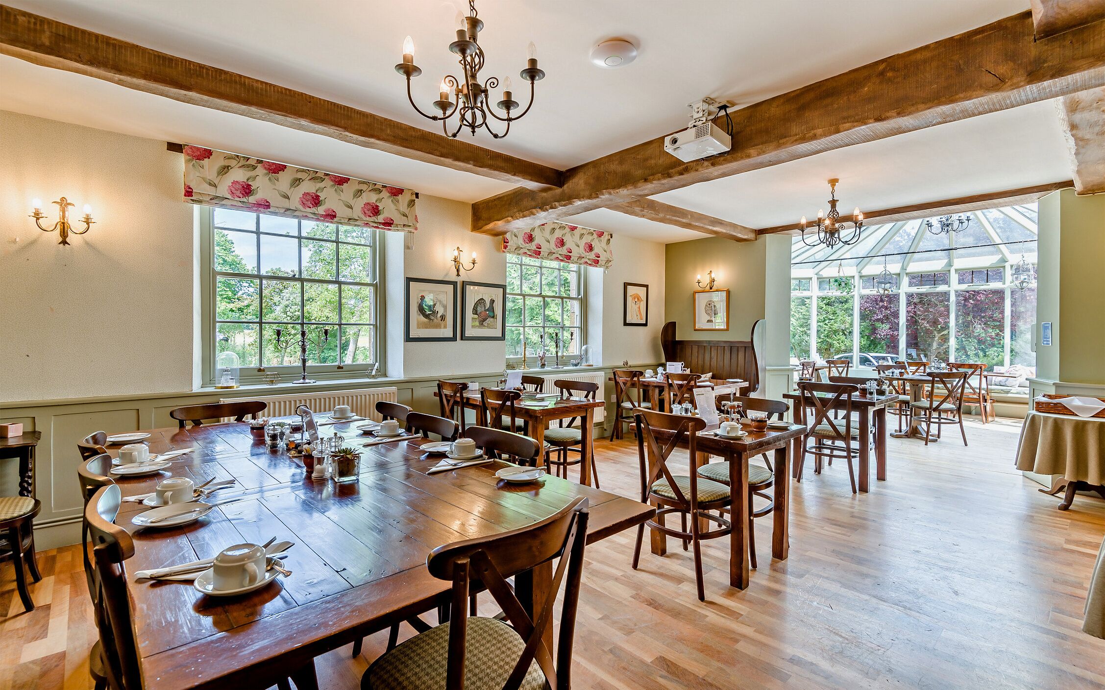 A spacious dining hall with wooden tables and chairs, large windows, and artwork on the walls.
