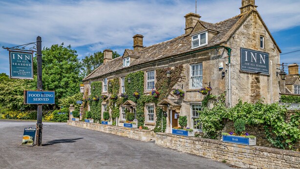 A traditional stone building with multiple signs indicating it is an inn offering a bar, restaurant, and hotel services. - Inn For All Seasons (Burford)