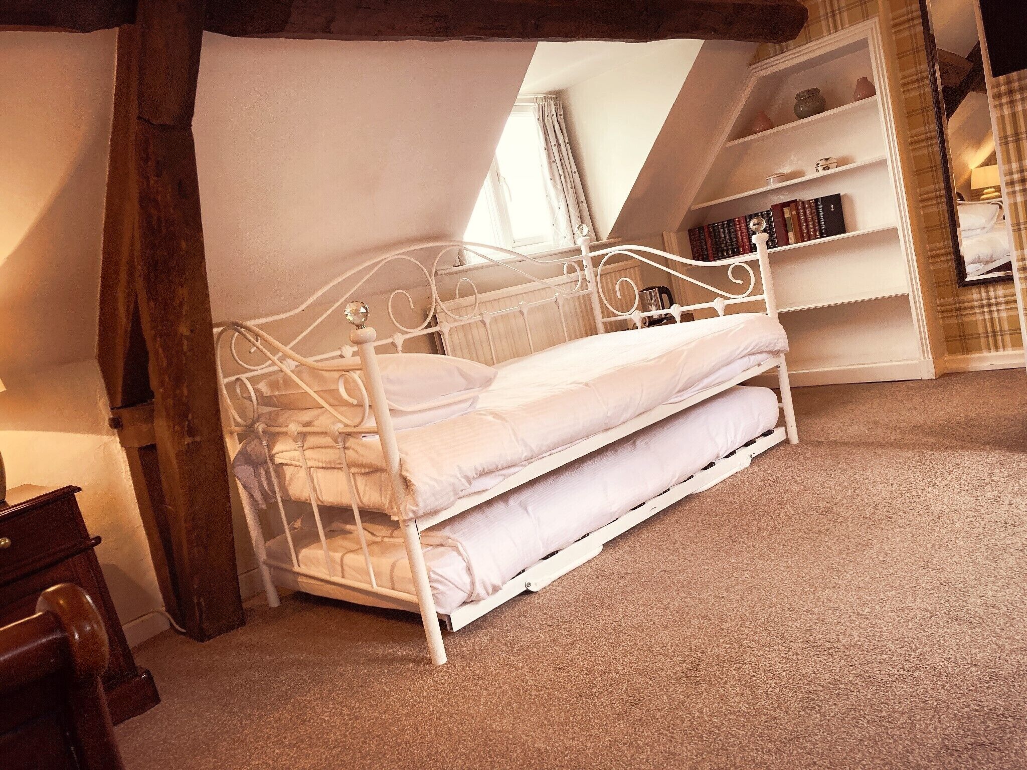 A white metal bed in a room with a sloped ceiling, a wooden chair, and a bookshelf with books.