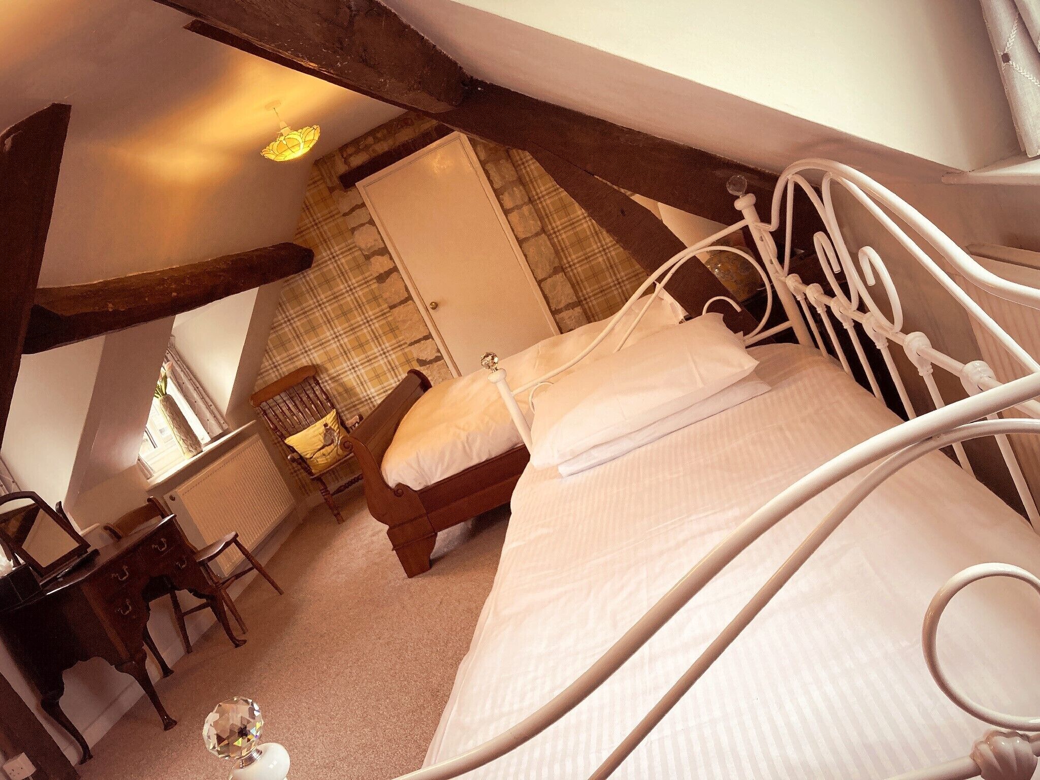 Une chambre avec un lit simple, une chaise en bois, un bureau et une fenêtre donnant sur une porte et un mur orné d’un motif à carreaux.