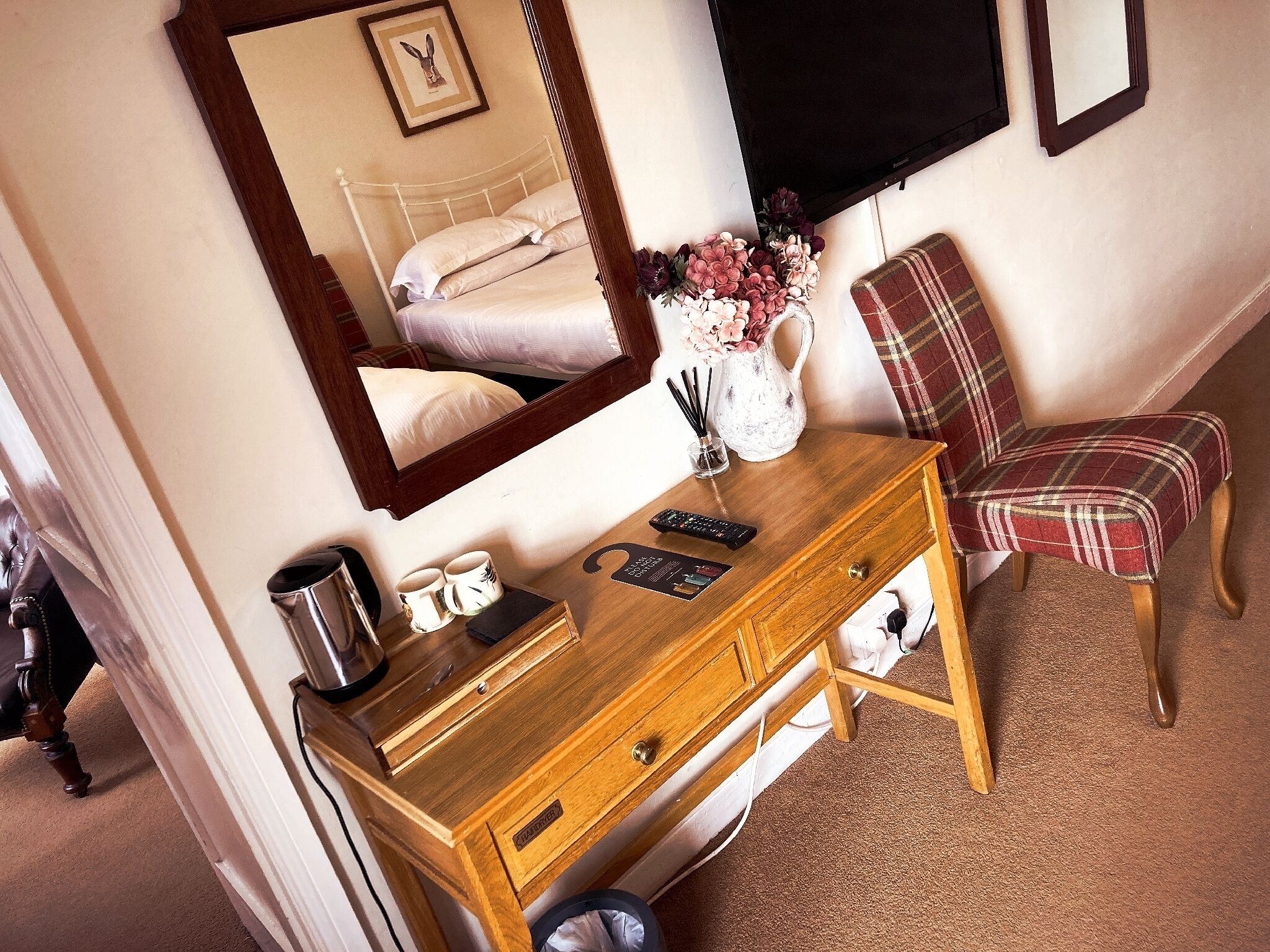 Ein Hotelzimmer mit einem Holzkasten, einem Spiegel, einem karierten Sessel, einer Vase mit Blumen und einem Bett mit weißen Laken.