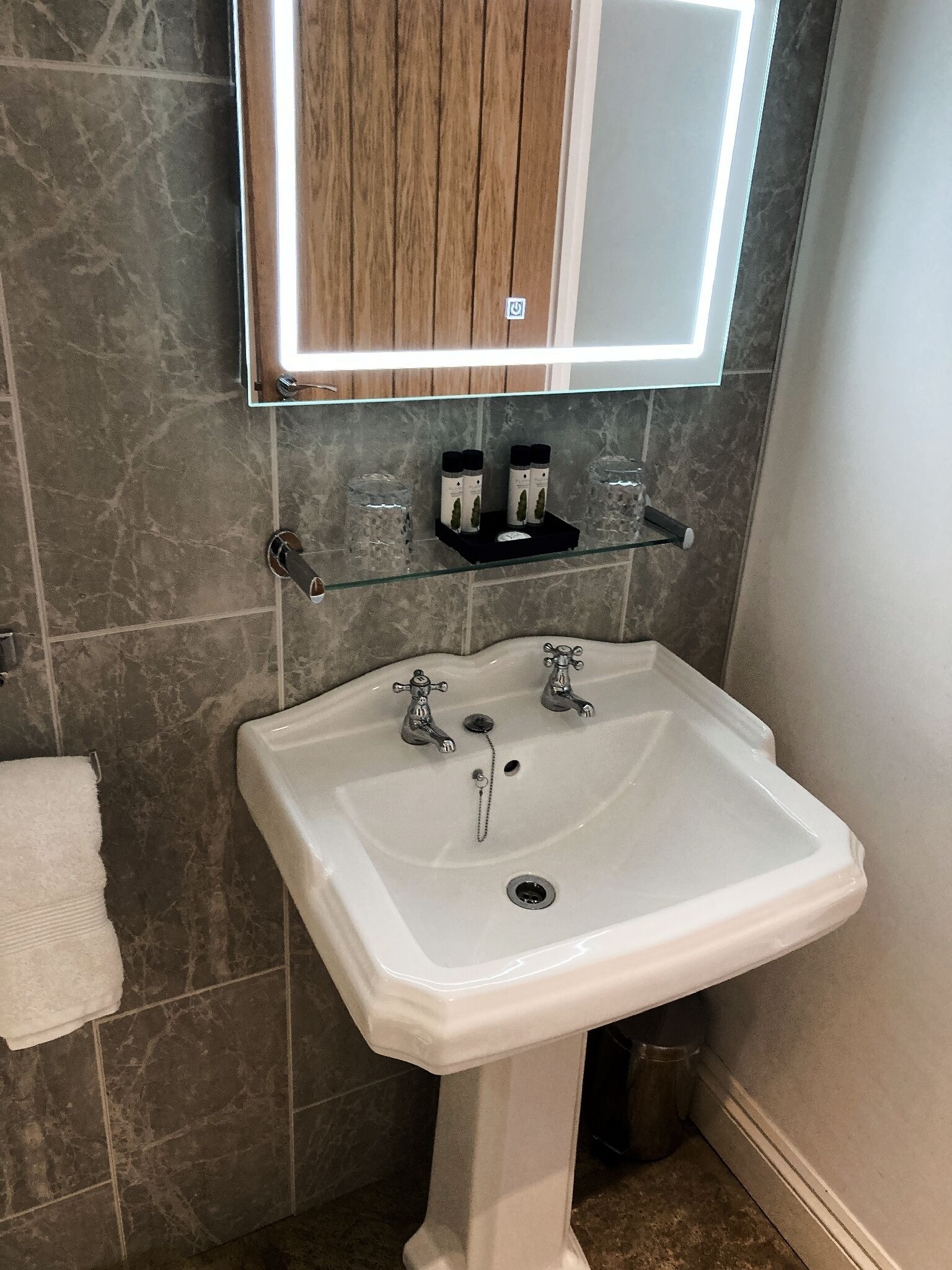 Casa de banho com lavatório pedestal branco, duas torneiras, espelho sobre o lavatório e prateleira com artigos de higiene pessoal.