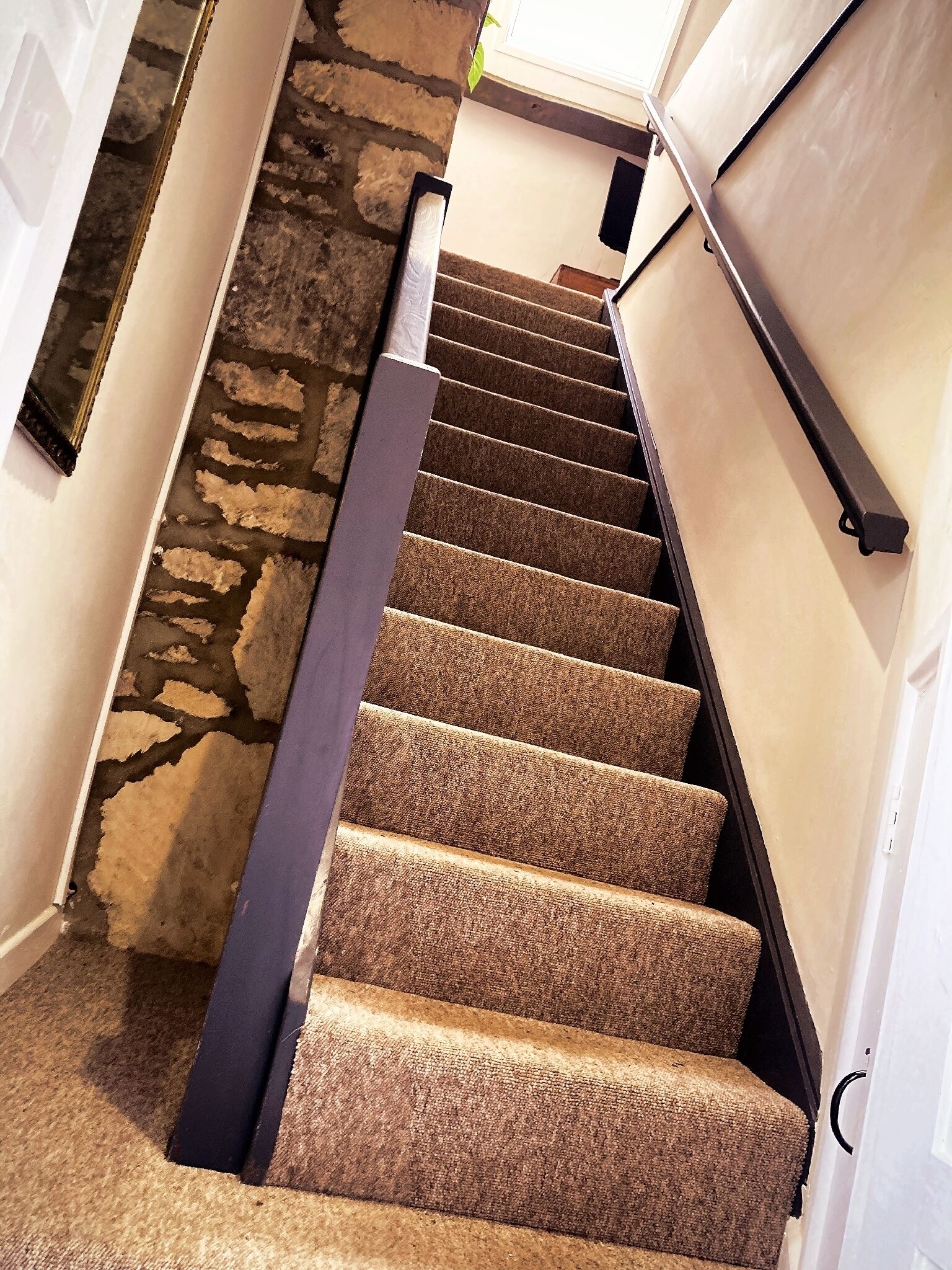 Una escalera alfombrada con barandilla que conduce a una habitación con pared de piedra y ventana.