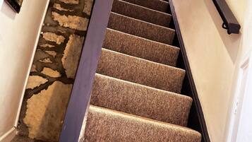 Una escalera alfombrada con barandilla que conduce a una habitación con pared de piedra y ventana.