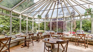 A conservatory with wooden furniture, large windows, and hanging plants. - Inn For All Seasons (Burford)
