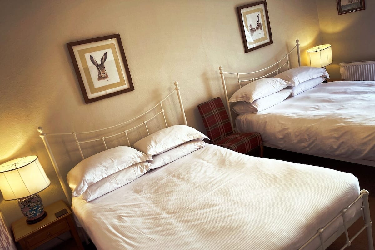 A hotel room with two beds, each with white linens and pillows, a plaid chair, and framed artwork on the walls.