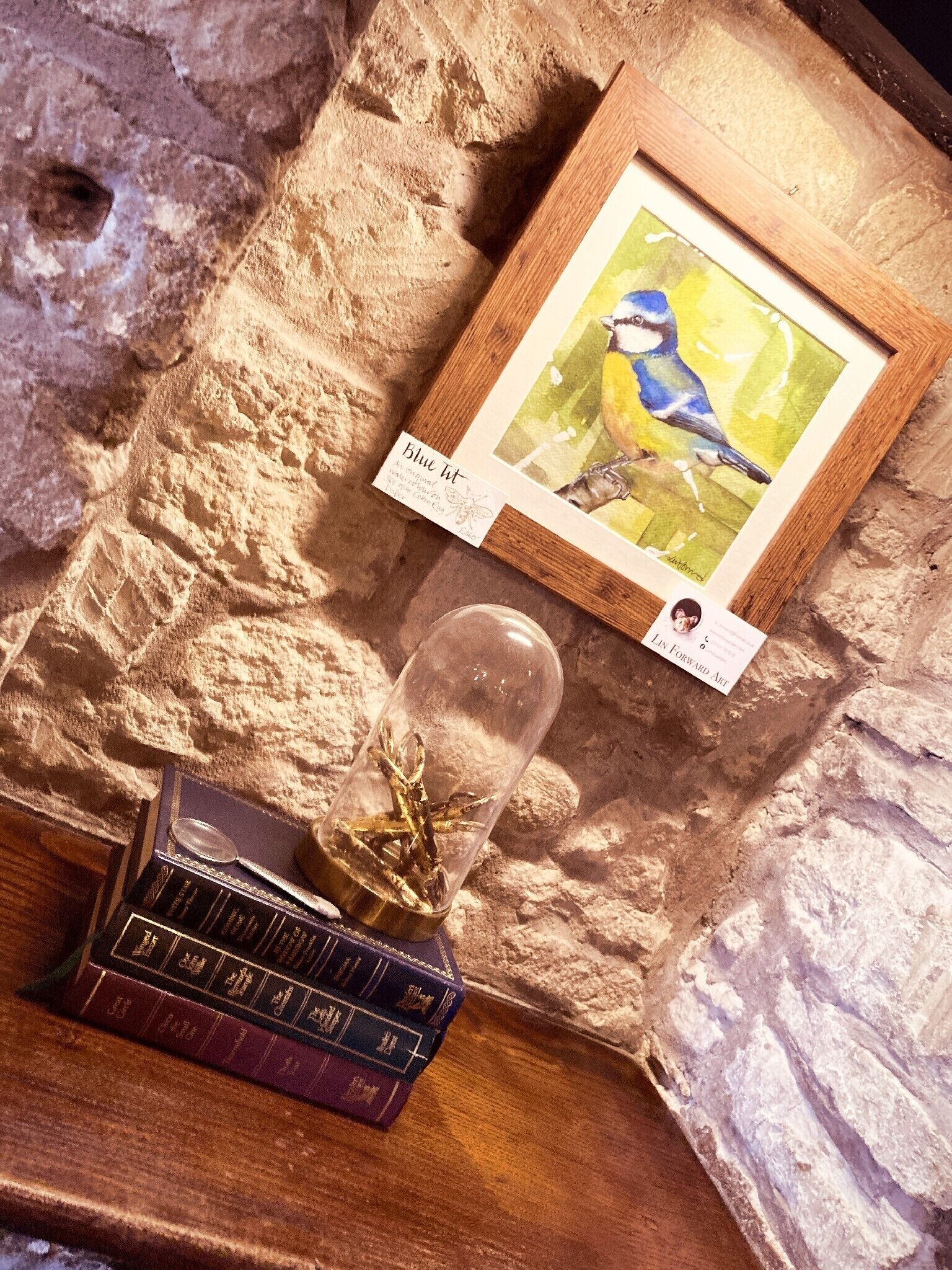 Una acuarela enmarcada de un pájaro, un terrario de vidrio con plantas, y una pila de libros sobre una pared de piedra texturizada.