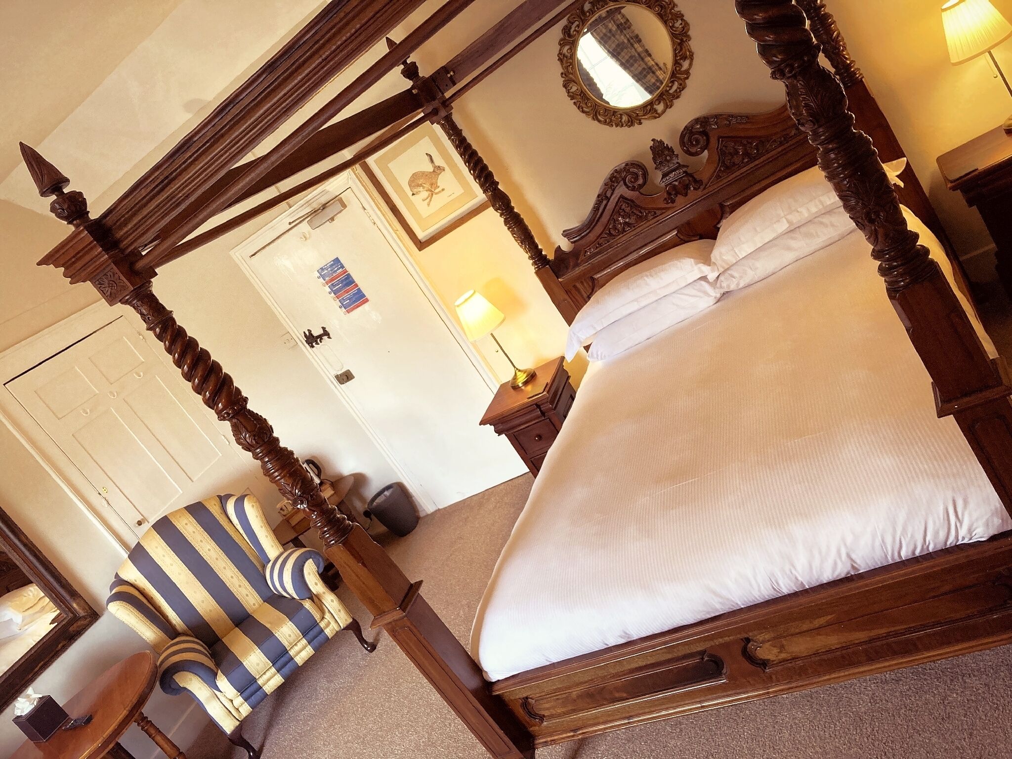 A four-poster bed with a wooden frame, a striped armchair, a bedside table with a lamp, and a framed picture on the wall.