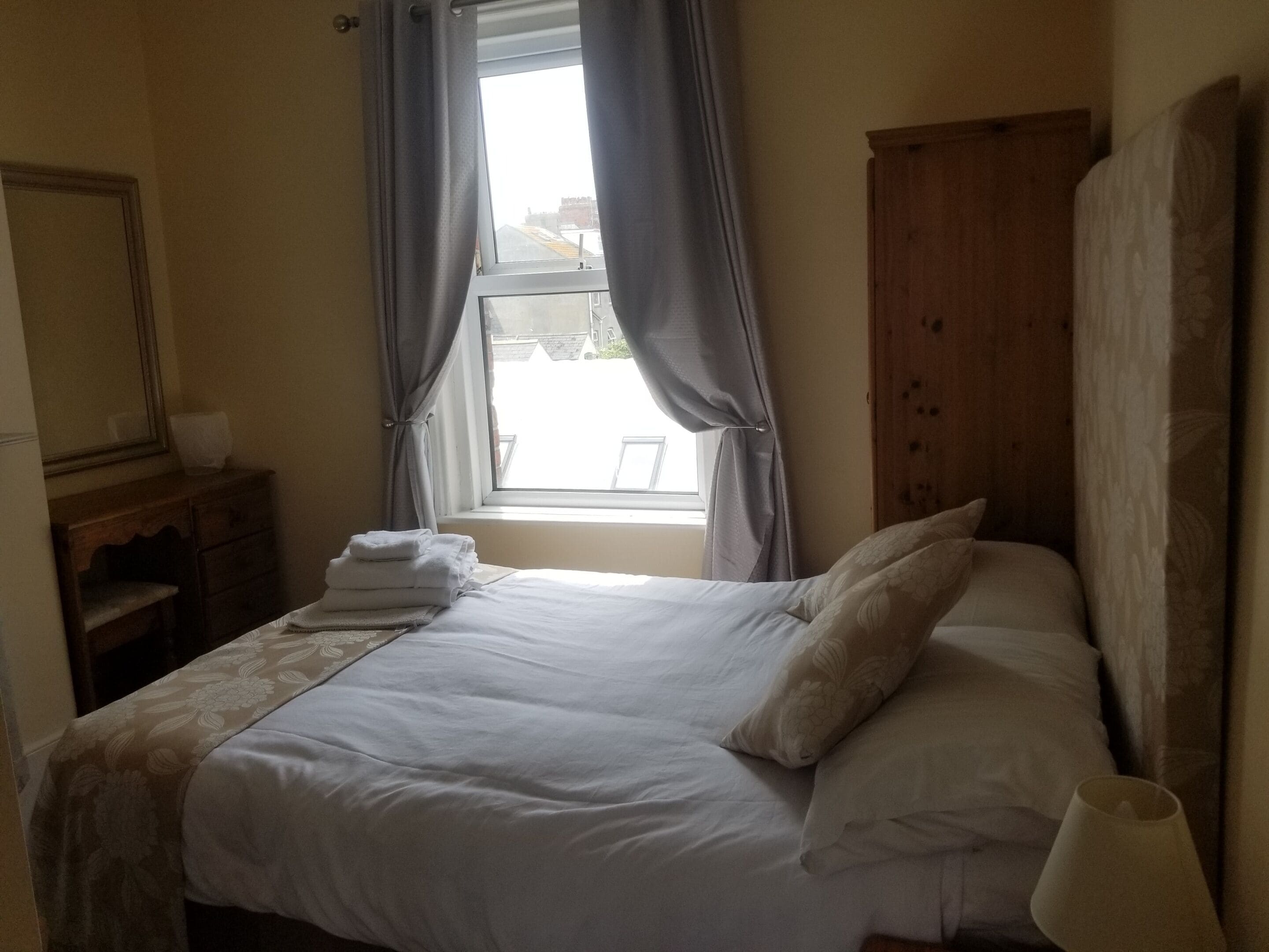 Ein ordentlich bezogenes Bett mit weißen Bettwäsche und einer zudecke mit Blumenmuster, ein Holzschrank, ein kleiner Nachttisch mit Lampe und ein Fenster mit Vorhängen.