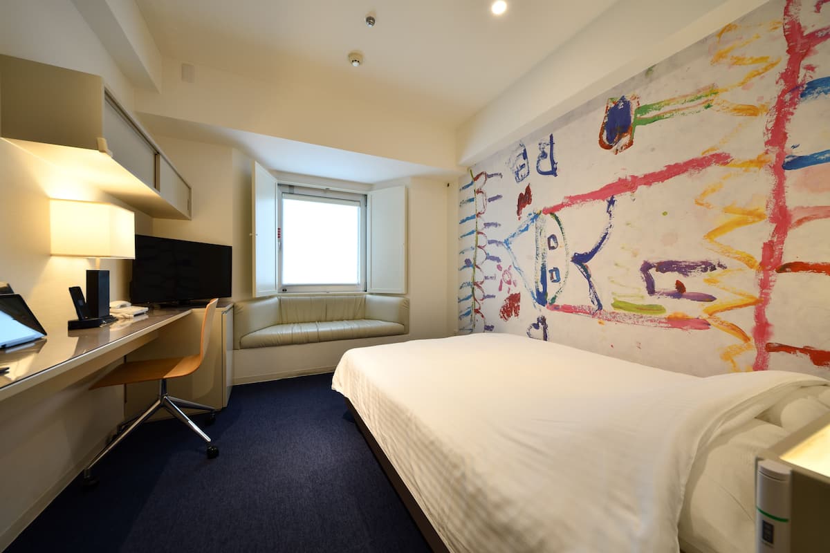 Une chambre d’hôtel avec un lit, un bureau, une chaise et une peinture abstraite aux couleurs vives accrochée au mur.