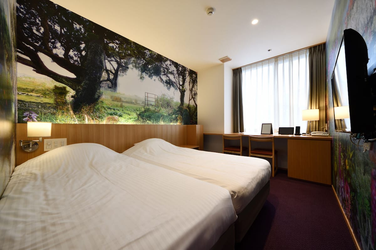 Une chambre d’hôtel avec deux lits, un bureau, une chaise, une télévision et un grand tableau au mur.
