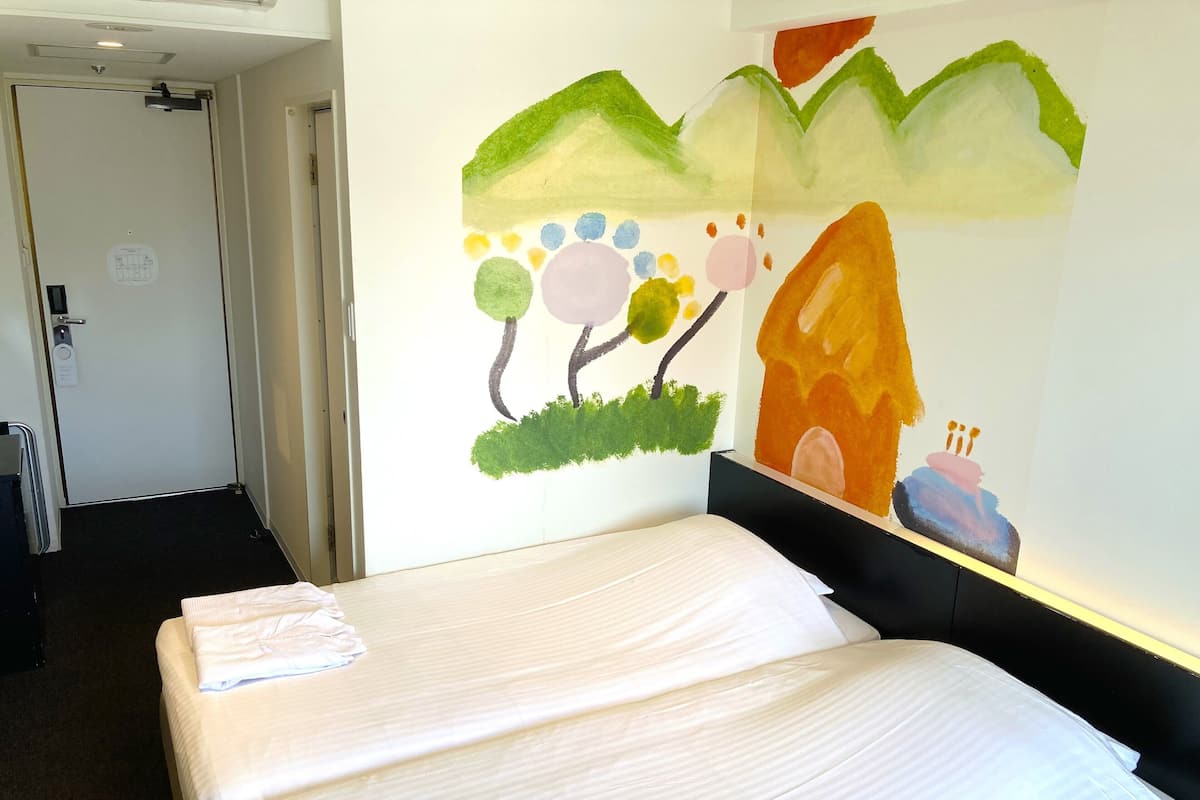 Une chambre d’hôtel avec un lit bien fait, un mur orné d’une fresque colorée et une porte sur le côté gauche.