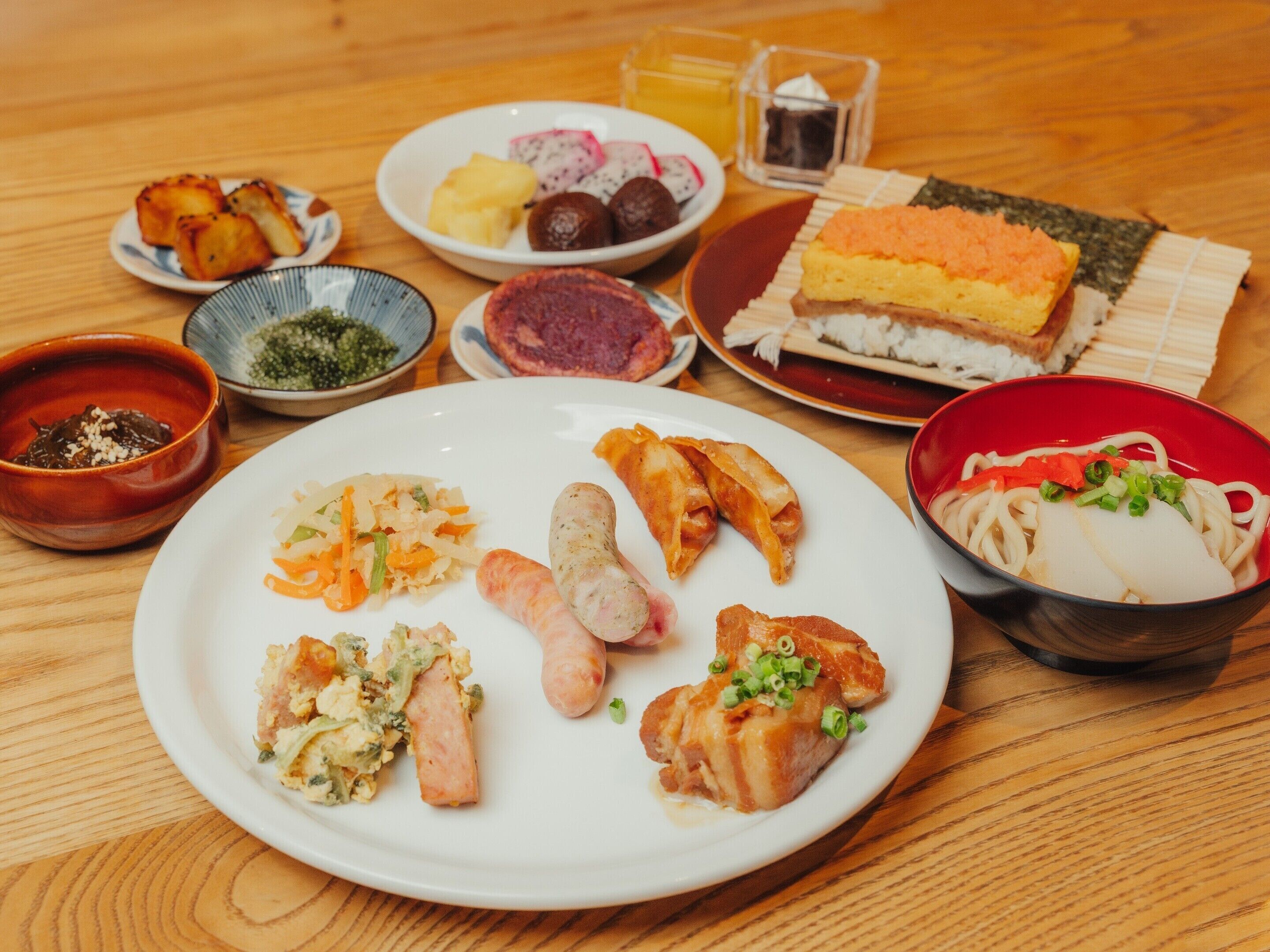 A variety of dishes on a wooden table, including a bowl of noodles, several plates with different foods, and a small glass of tea.