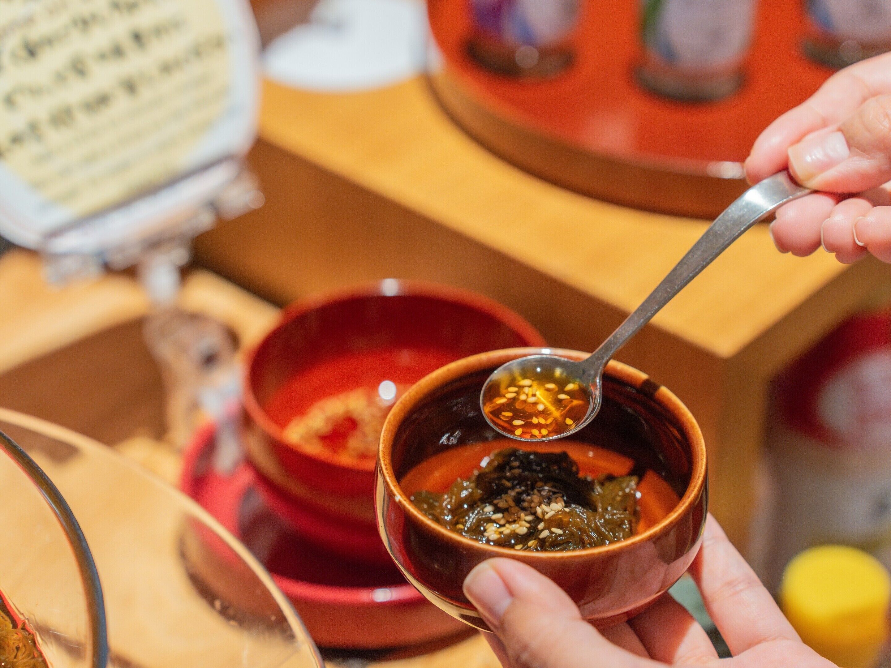 A person holding a small bowl with a spoon containing a dark liquid and sesame seeds.