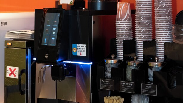 A coffee machine with a digital display, a menu board with coffee options, and stacked paper cups. - HOTEL ART STAY NAHA Kokusai Street (Naha)
