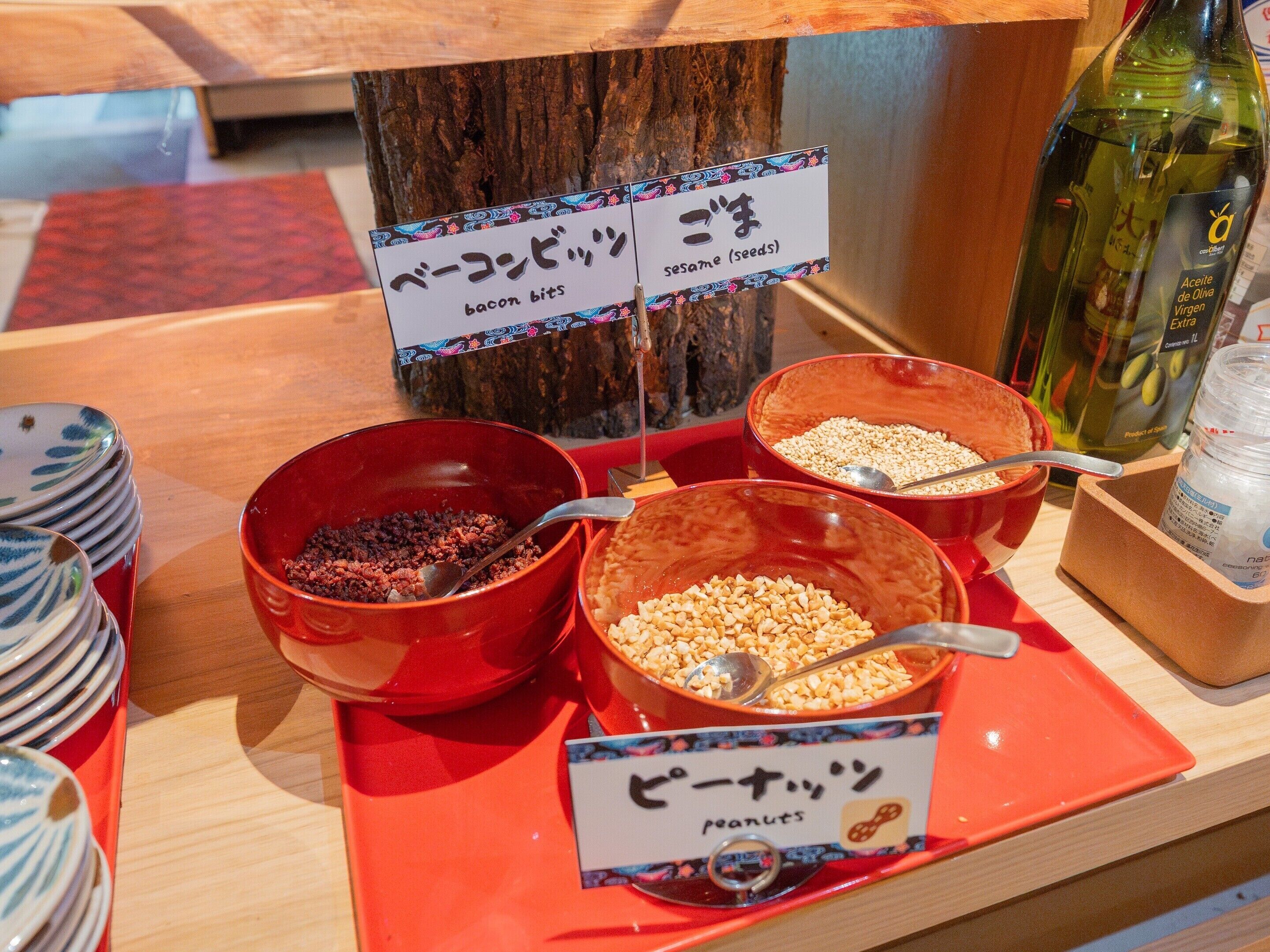 A buffet table with labeled bowls of bacon bits, sesame seeds, and peanuts.