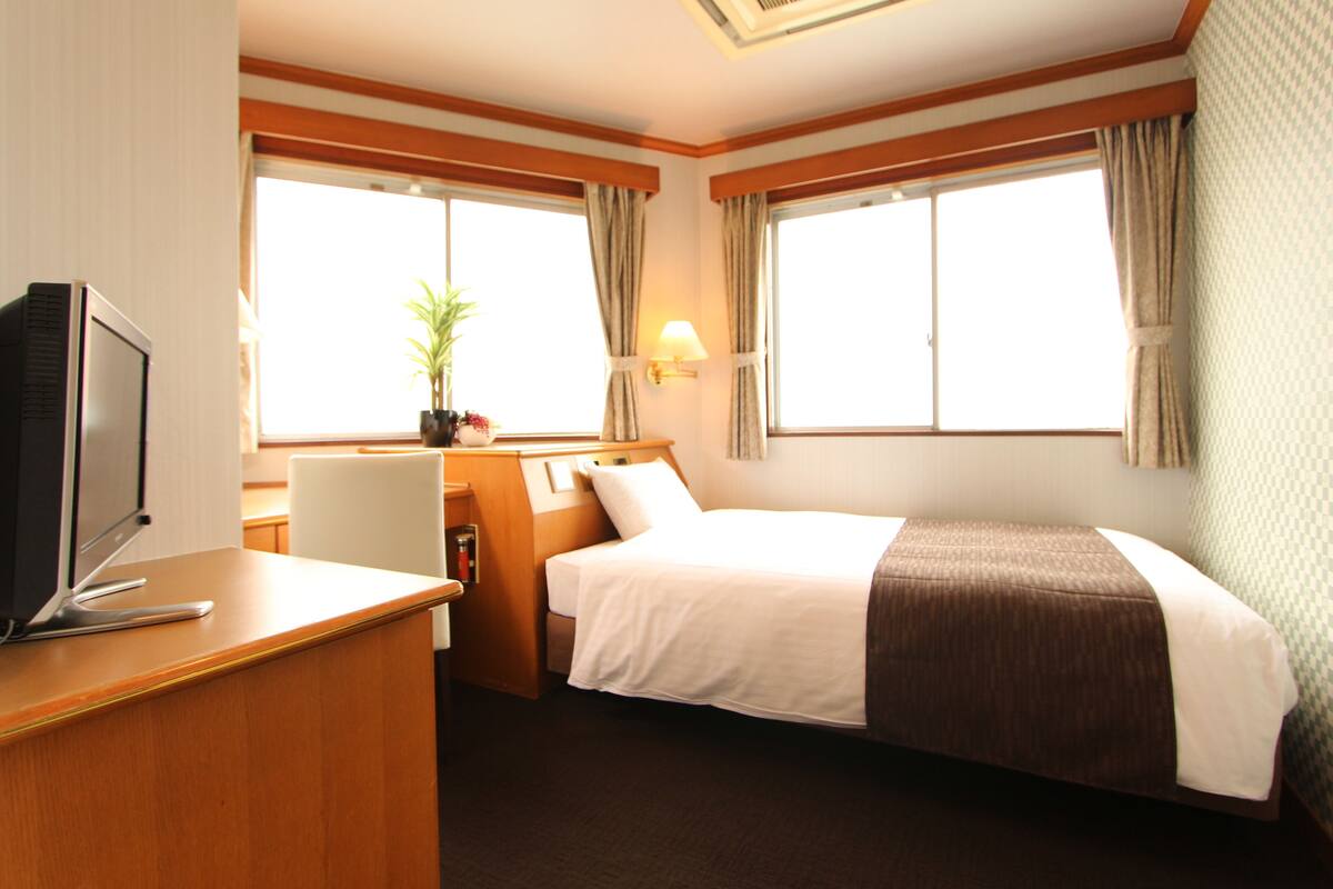 A hotel room with a bed, a desk with a computer, a chair, a TV, and a window with curtains.