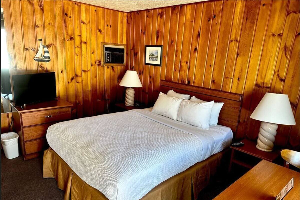 A wooden-paneled room with a bed, two bedside lamps, a TV on a wooden dresser, and a small trash bin.