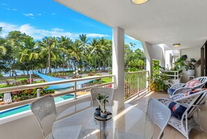 Uma varanda com vista para uma piscina, palmeiras e cadeiras de descanso.