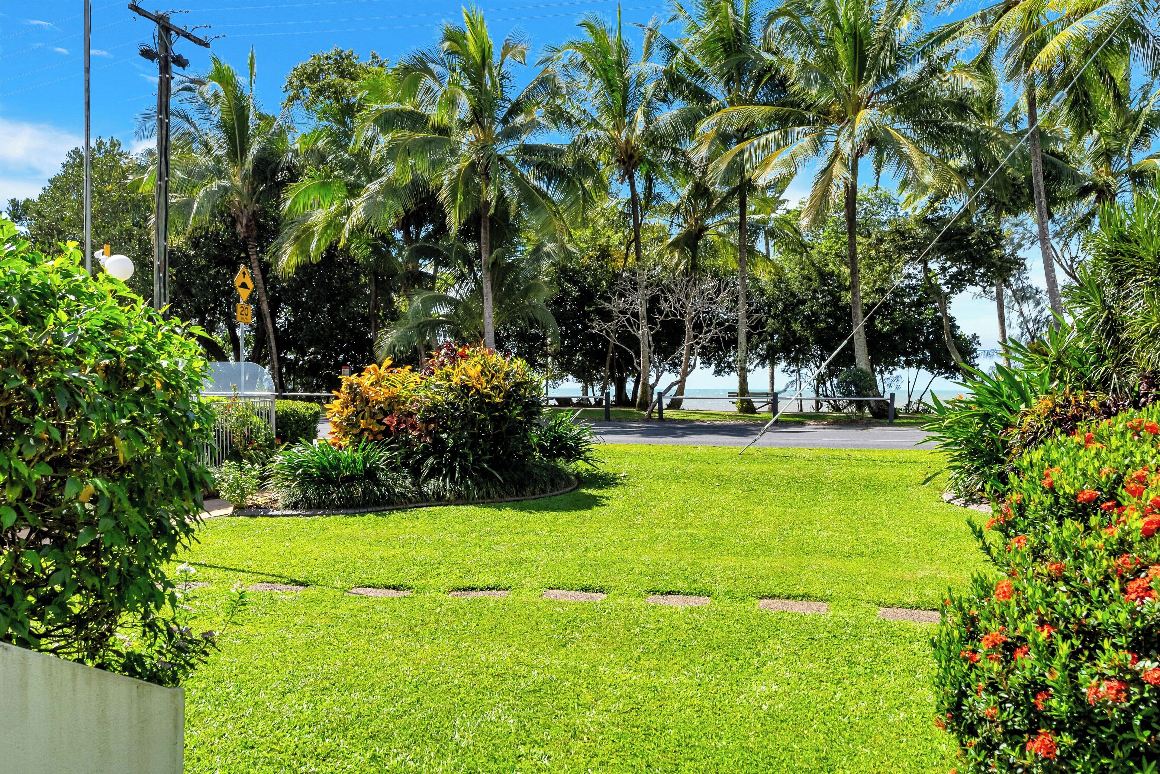 A well-maintained garden with a paved pathway, palm trees, and a view of the ocean.