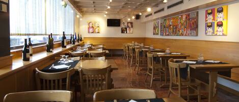 Interior de restaurante con mesas y sillas de madera, una vitrina de vinos y obras de arte coloridas en las paredes.