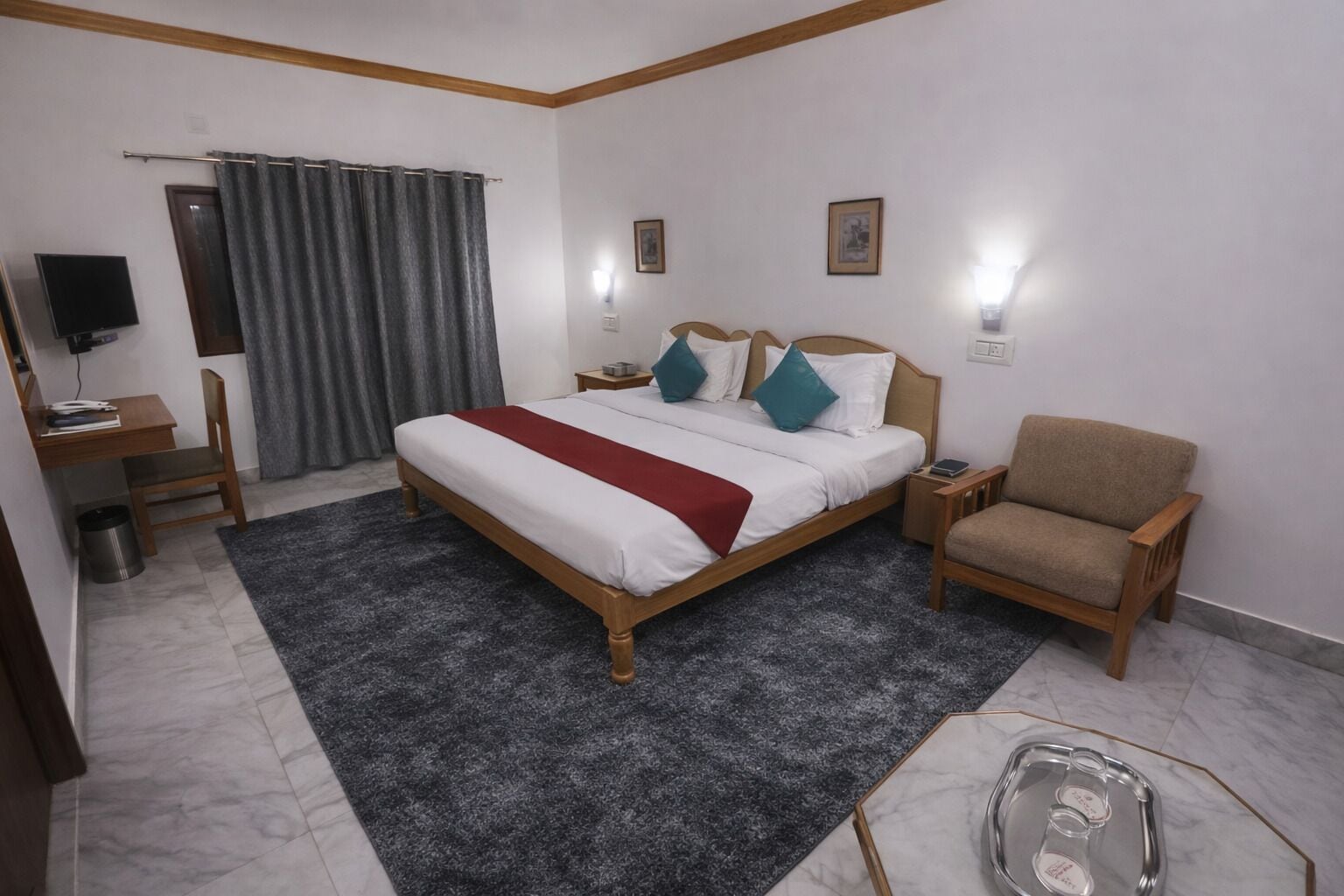 Quarto de hotel com cama, poltrona, escrivaninha, televisão e janela com cortinas.