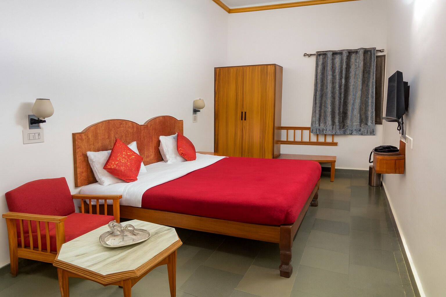 A hotel room with a wooden bed, red bedding, a wooden wardrobe, a small table with a vase, and a TV on the wall.