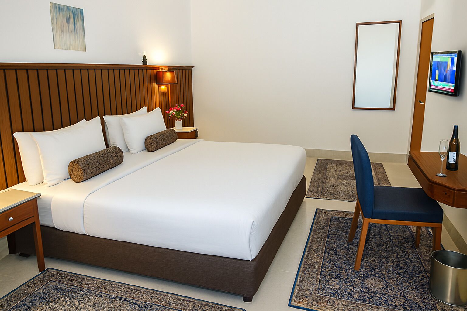 Habitación de hotel con una cama grande, cabecera de madera, mesita de noche, escritorio con silla, televisor y un cesto de basura pequeño.