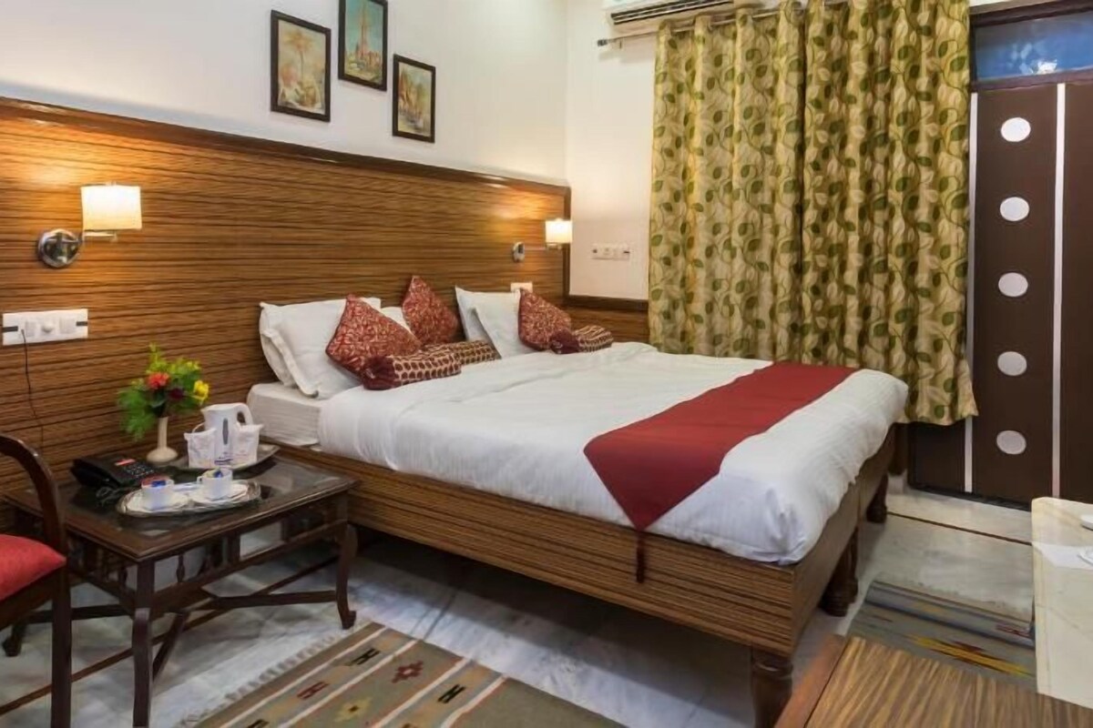 A hotel room with a wooden headboard, a bed with a red blanket, a small table with a vase of flowers, and a chair.