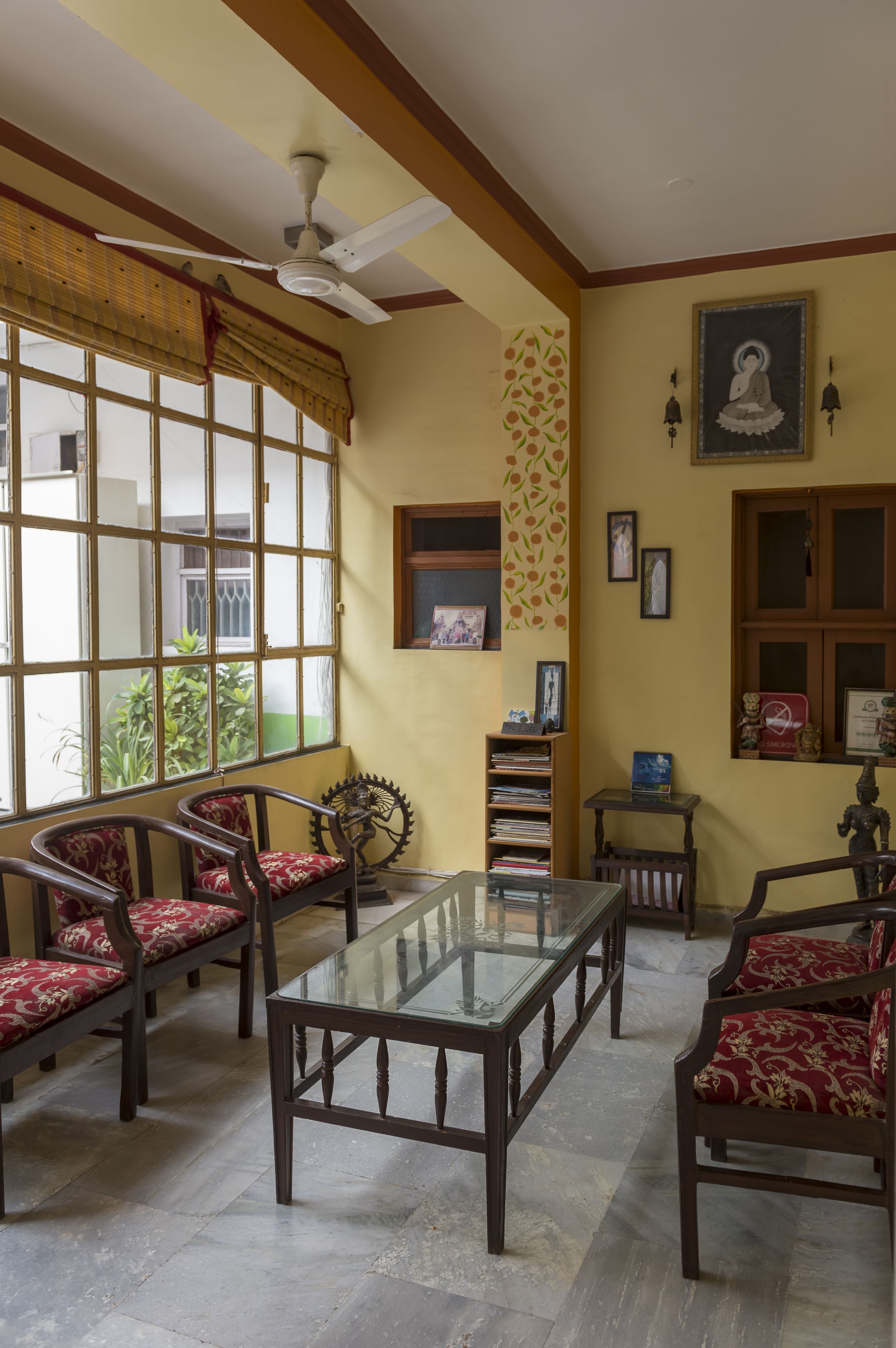 A room with a glass-top table, wooden chairs with red cushions, a ceiling fan, and a window with a view of greenery outside.