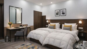 A hotel room with a large bed, a desk, a chair, a small table with a tray of cups and a pitcher, and a trash can. - Hotel Harmony (Khajuraho)